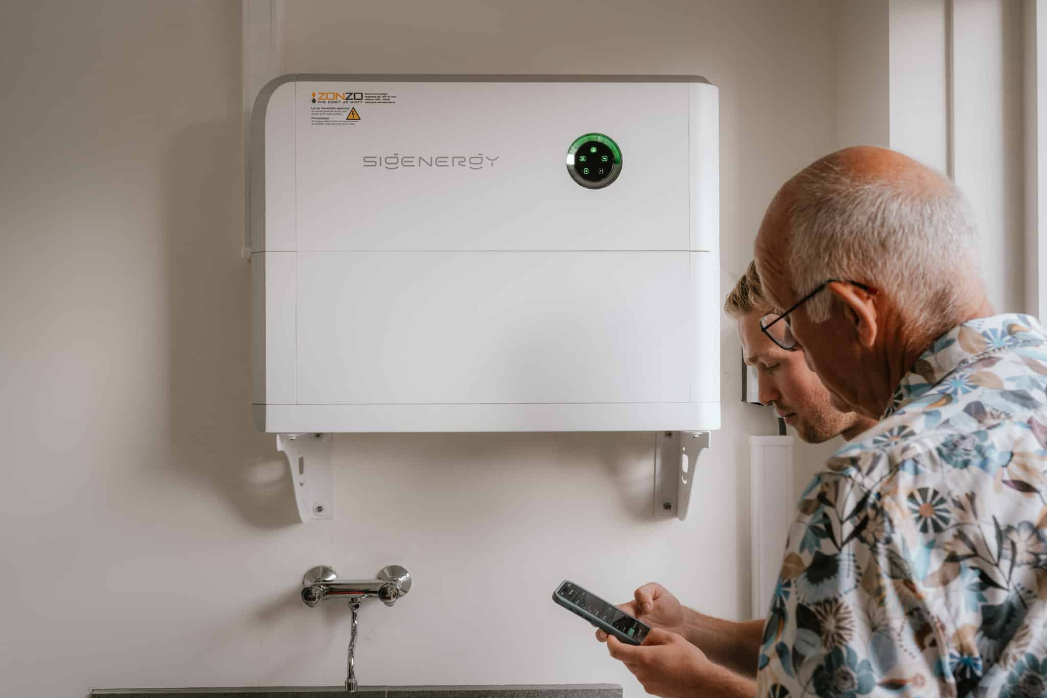 Thuisbatterij. Sigenergy Thuisbatterij. Thuisbatterij hangend aan de muur. Rechterzijde een medewerker van Zonzo die uitleg geeft aan een klant over de werking van de batterij en het bijbehorende overzicht op een telefoon
